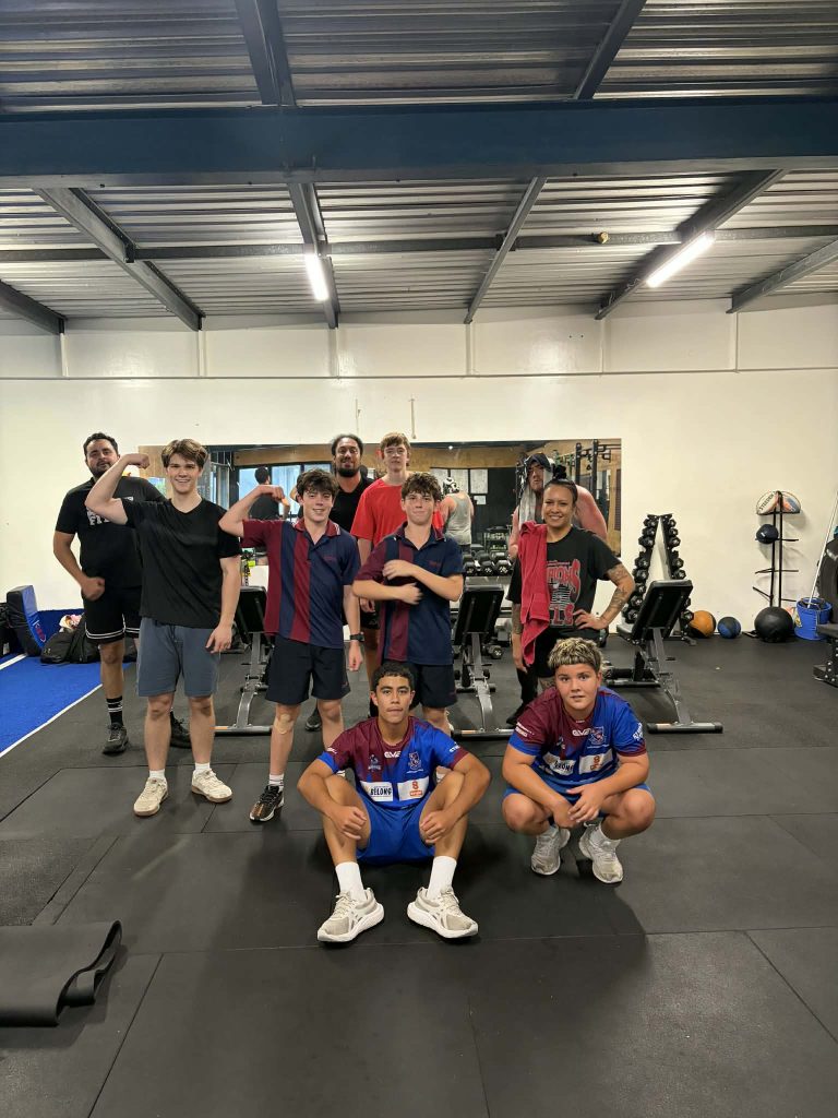 Group of young people in a gym posing for a photo, some flexing their arms in front of weight machines and mirrors.