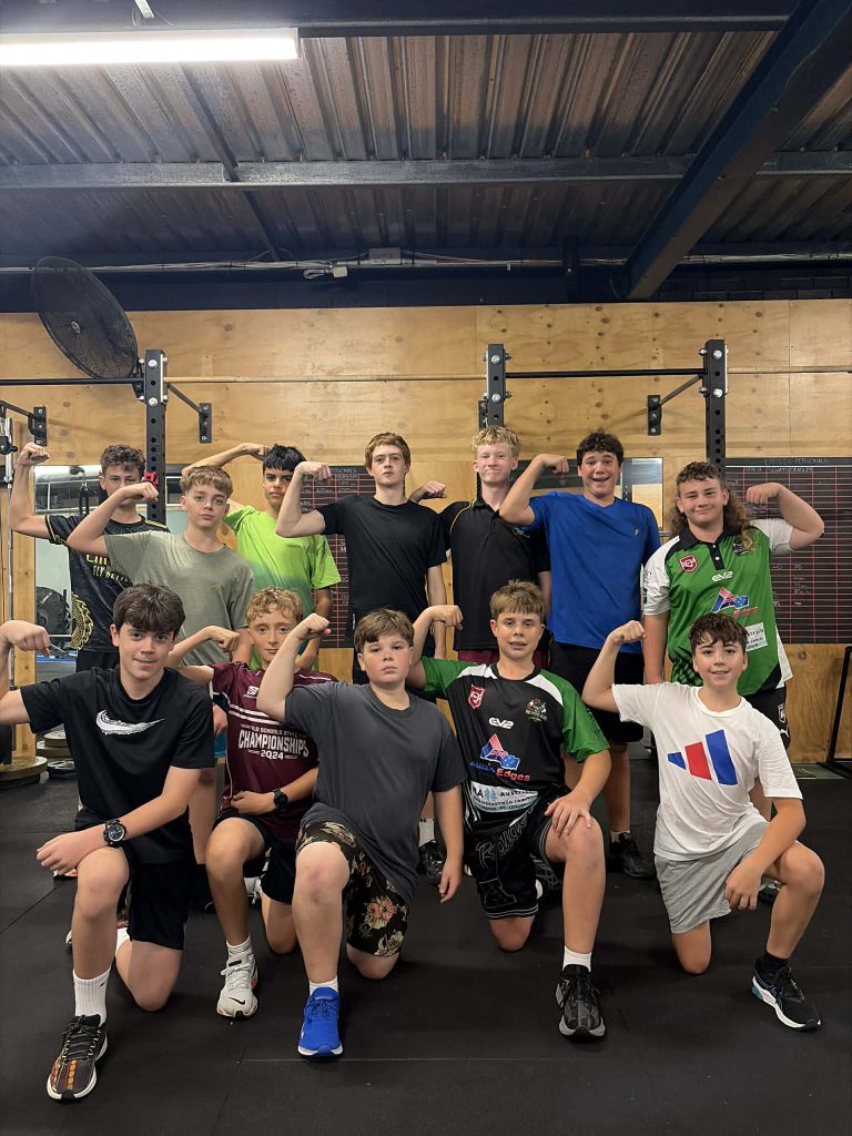 Group of young athletes posing and flexing muscles in a gym, wearing workout clothes and kneeled in front, with a wooden wall and gym equipment behind them.